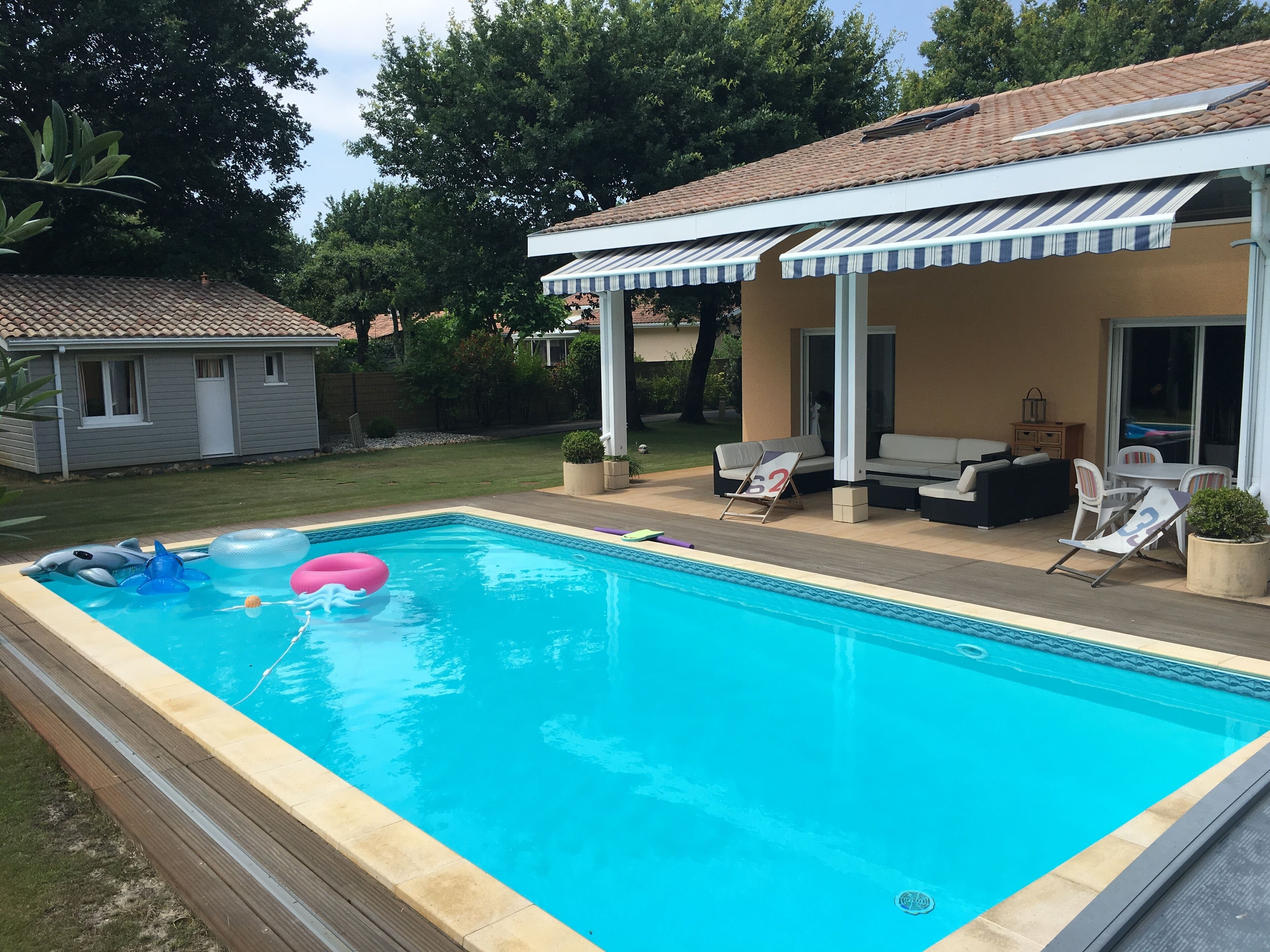 Ein rechteckiger Pool mit gefliestem Deck, eine überdachte Terrasse mit Sitzgelegenheiten und ein kleines Haus im Hintergrund.