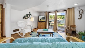 A room with a bed, a television, a white wicker chair, a wooden table, and a view of a green landscape through the door.