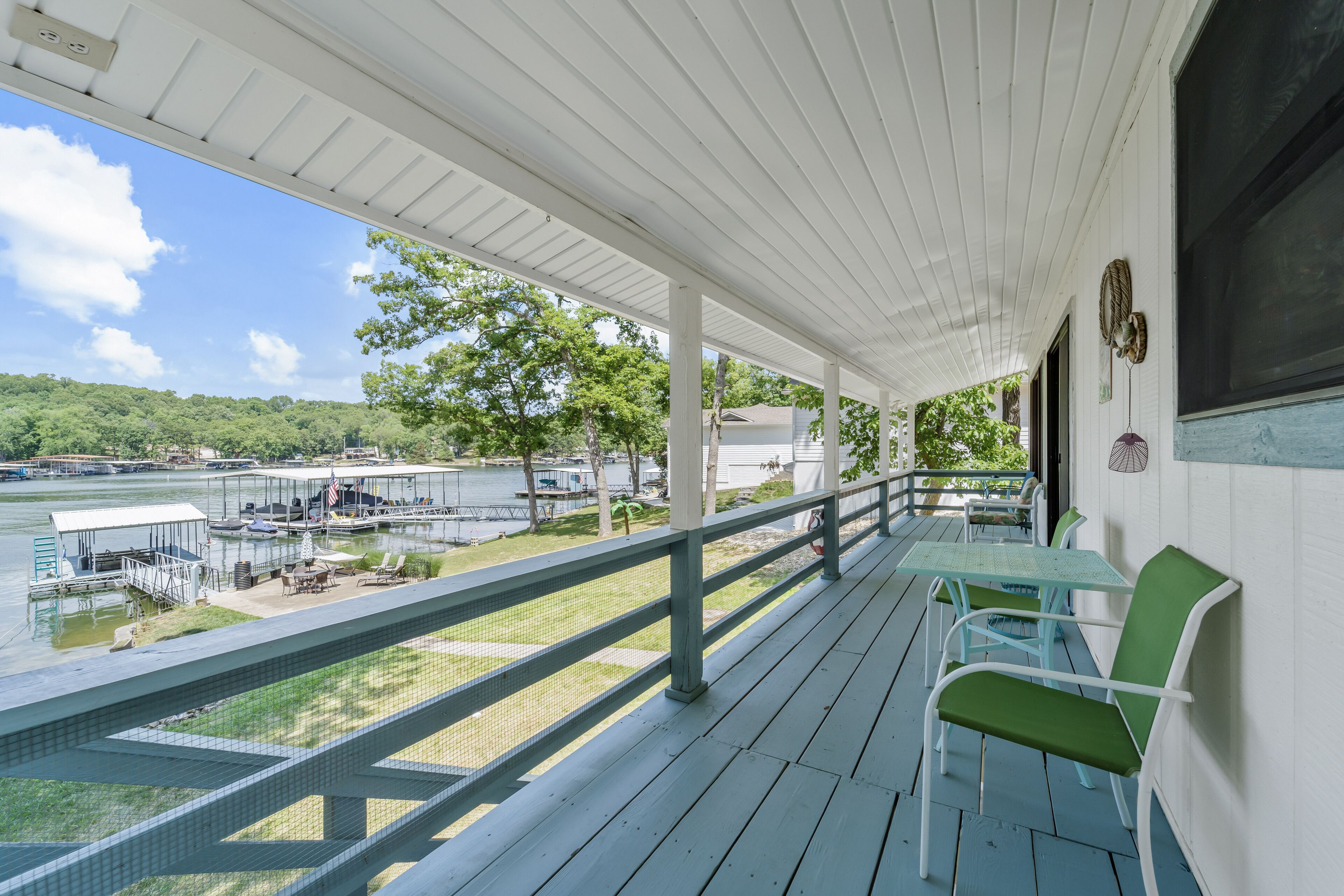 Une terrasse extérieure couverte, avec vue sur un lac et une zone de quai, agrémentée de chaises vertes et d’une table.