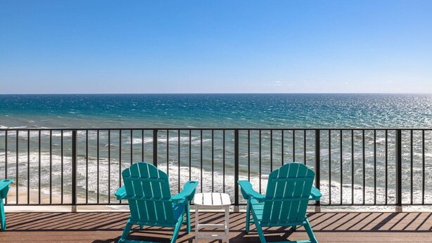Una terrazza con due poltrone Adirondack verdi e un tavolino, affacciata su una spiaggia con acqua cristallina e un corrimano in metallo.