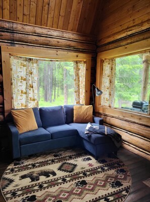 Un intérieur de chalet chaleureux avec un canapé d’angle bleu, un tapis à motifs et de grandes fenêtres avec des rideaux.