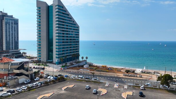 A modern hotel with a unique triangular architecture, a parking lot, and a view of the sea with sailboats.