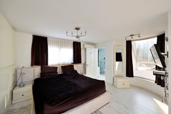 A bedroom with a large bed, bedside tables, a nightstand, a mirror, a window with curtains, and a ceiling light fixture.