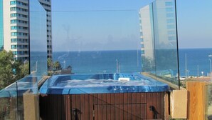 A hot tub with a wooden deck and glass panels, overlooking a body of water and buildings.