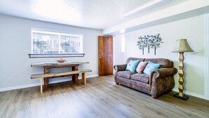Uma sala de estar com um banco de madeira, um sofá marrom com almofadas, piso de madeira, uma janela com persianas e um adorno na parede.