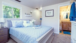 Quarto com cama, mesa de cabeceira, penteadeira, janela e banheiro visível através de uma porta aberta.