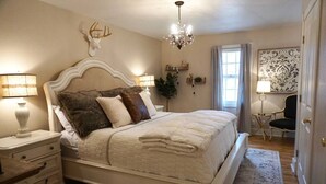 Een slaapkamer met een groot bed, nachtlampen, een kroonluchter, een hertenkop aan de muur, en een raam met gordijnen.