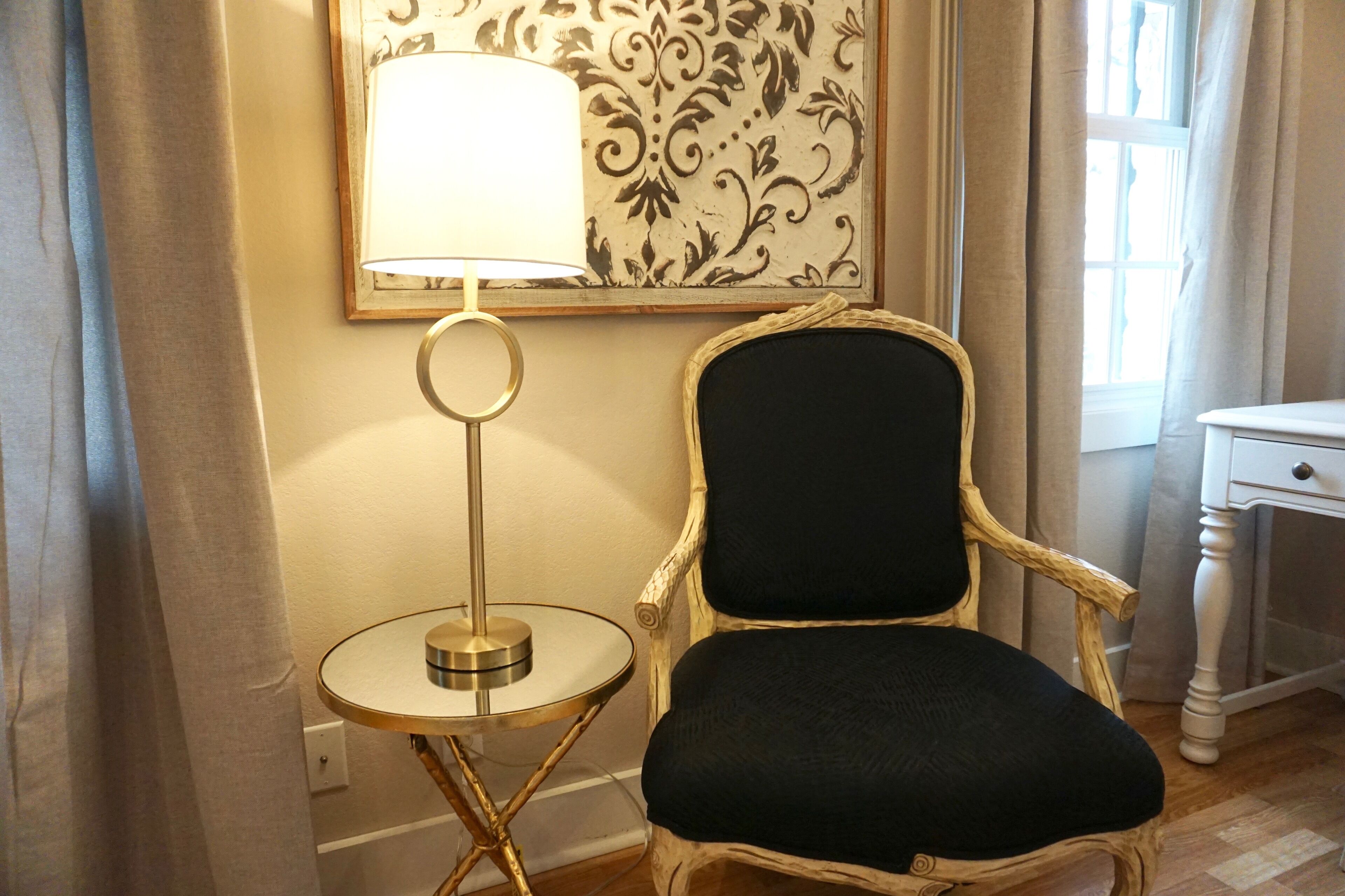 Une pièce avec une table ronde, une chaise, une lampe, une petite table blanche et une fenêtre avec des rideaux.