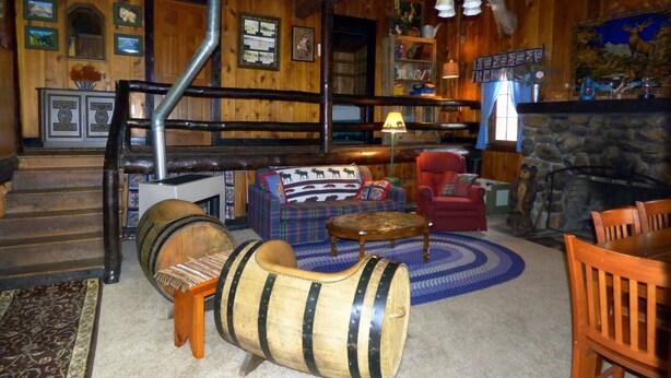 A rustic interior with wooden walls, a stone fireplace, a ceiling fan, and a cozy seating area with a plaid sofa and armchairs. - Pine Crest- Historic 3 bedroom Log Cabin built in the 1930's by the CCC.   (Custer)