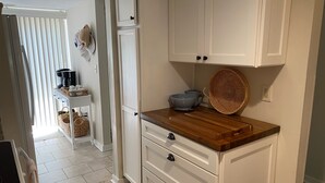 Uma cozinha com armários brancos, bancada de madeira e uma cafeteira. Há uma cesta sobre a bancada e uma cesta de palha na parede.