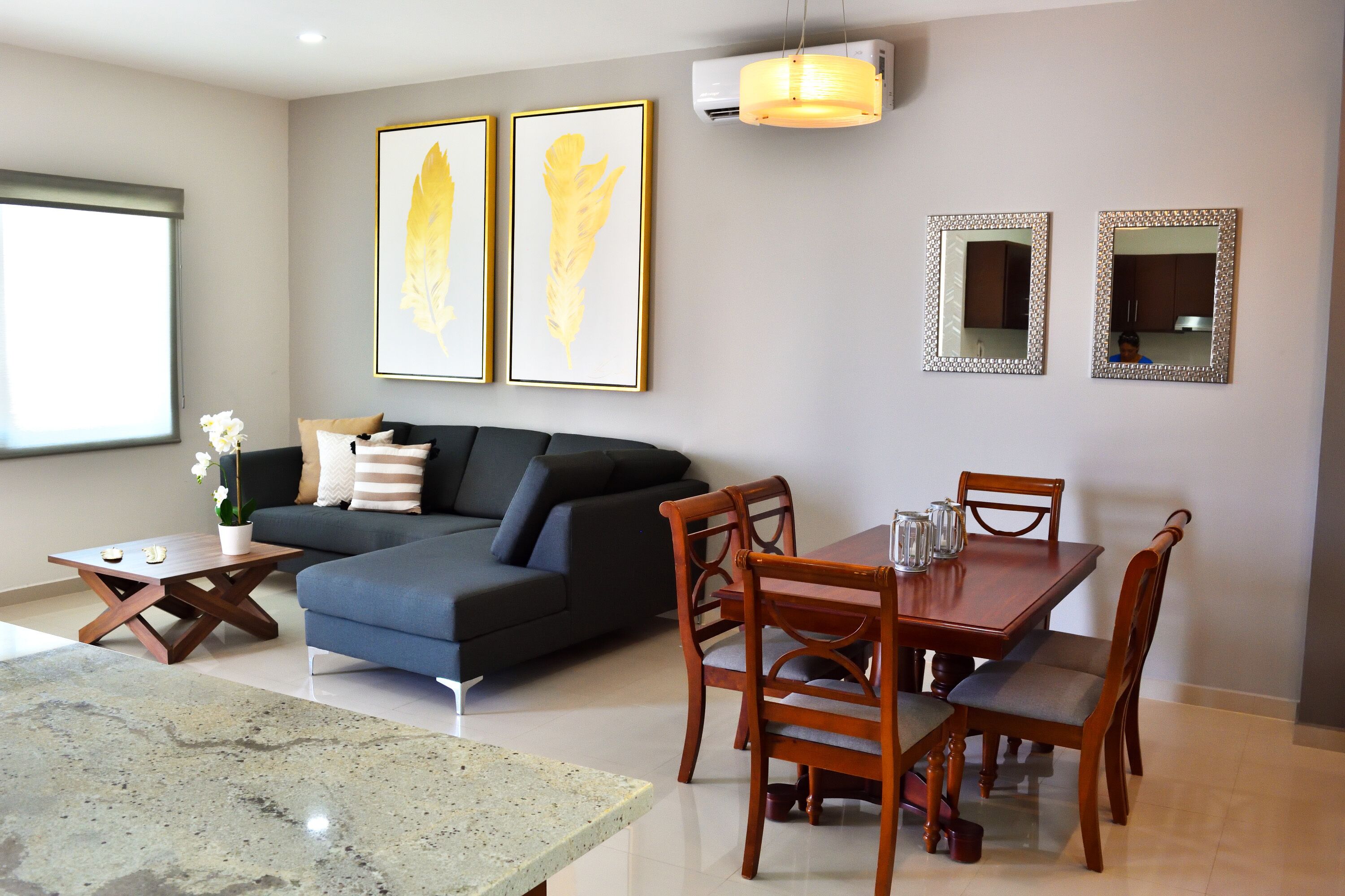 A modern living room with a dark blue sofa, wooden dining table, and chairs. There are two framed artworks on the wall and an air conditioning unit above the sofa.