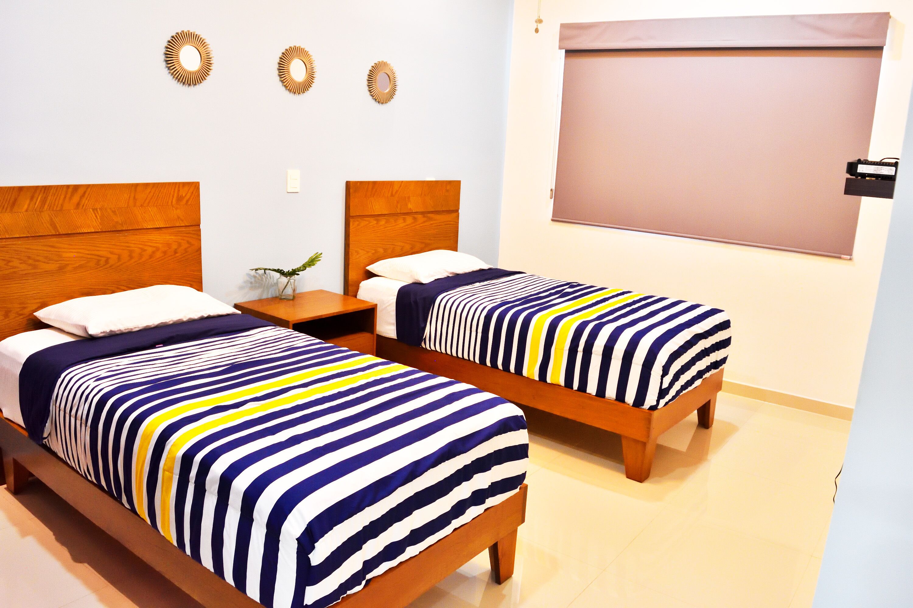 Una habitación de hotel con dos camas de madera, cada una con colcha a rayas, una mesita de noche con un florero y una ventana con persiana.