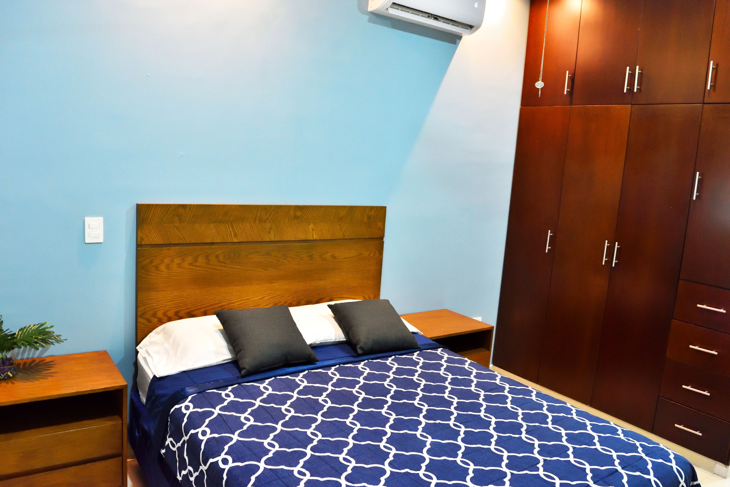 Une chambre avec une tête de lit en bois, un lit recouvert d’une couvre-lit à motifs, une table de chevet, une armoire et un appareil de climatisation fixé au mur.