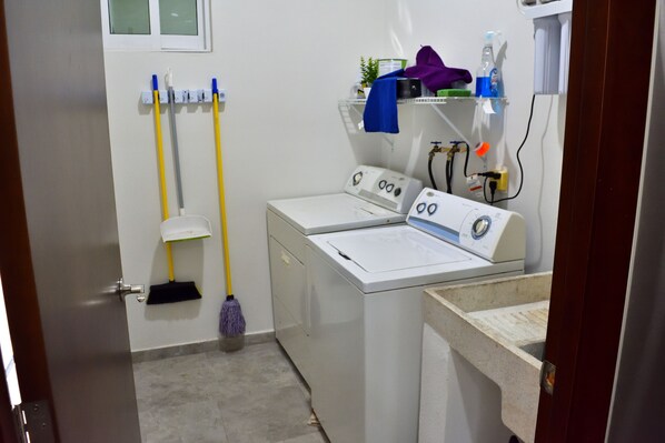 A laundry room with two washing machines, a mopping bucket, and a shelf with cleaning supplies. - Ocean View, Steps from Beach... Brand New (Mazatlán)