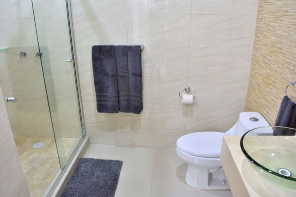 A modern bathroom with a glass shower enclosure, a white toilet, a glass sink, and a beige tiled wall. - Ocean View, Steps from Beach... Brand New (Mazatlán)