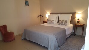 Une chambre à coucher bien rangée, avec un grand lit, deux tables de chevet avec des lampes, un fauteuil rouge et une petite table sur laquelle se trouve un vase et une rose.