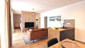 Ein modernes Wohnzimmer mit Flachbildfernseher, braunem Ledersofa, Esstisch aus Holz mit Obstschale und einer Küchenzeile mit Spüle und Arbeitsplatte.