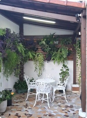 An outdoor patio with a tiled floor, white metal chairs, and a variety of potted plants.