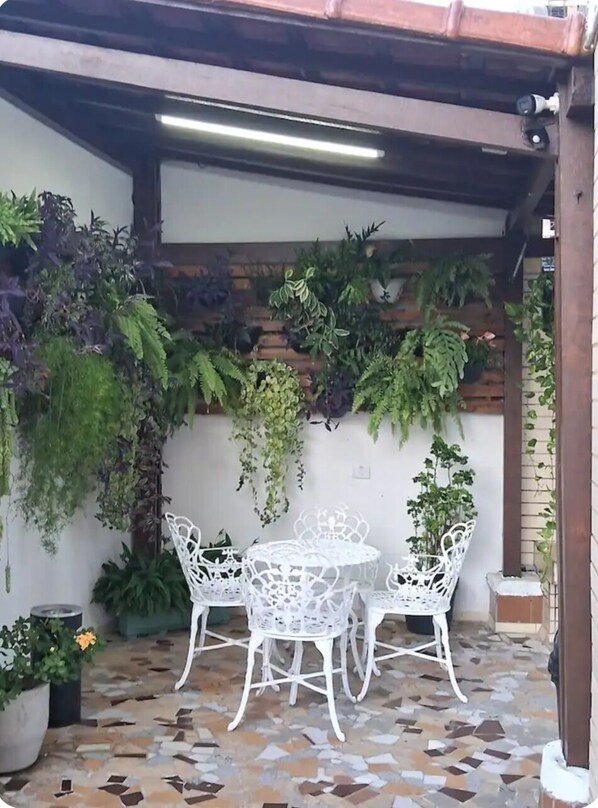 An outdoor patio with a tiled floor, white metal chairs, and a variety of potted plants.