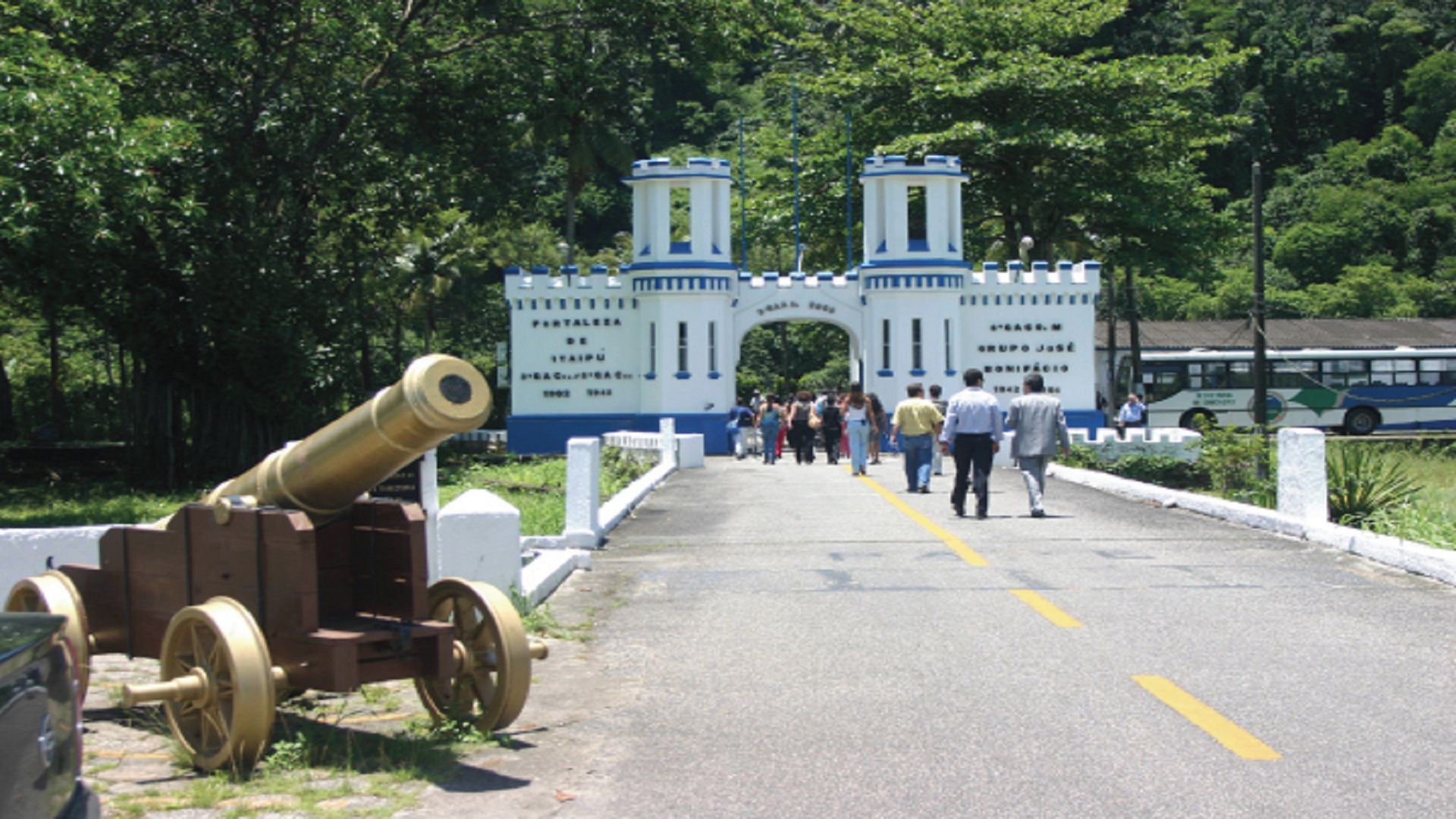A historical cannon on a paved road with a white and blue gate structure in the background.