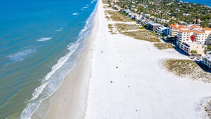 Vue aérienne d’un hôtel situé en bord de mer, avec une longue plage, des eaux d’un bleu limpide et une zone résidentielle dense en arrière-plan.