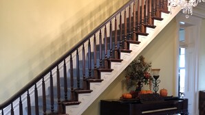 A grand staircase with wooden balusters and a chandelier, leading to a room with a grand piano and a decorative vase. - Loyd Hall Plantation (Cheneyville)