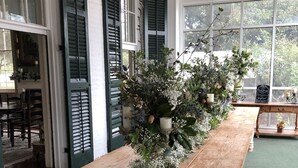 A long wooden table with a floral arrangement in a room with large windows and green shutters. - Loyd Hall Plantation (Cheneyville)