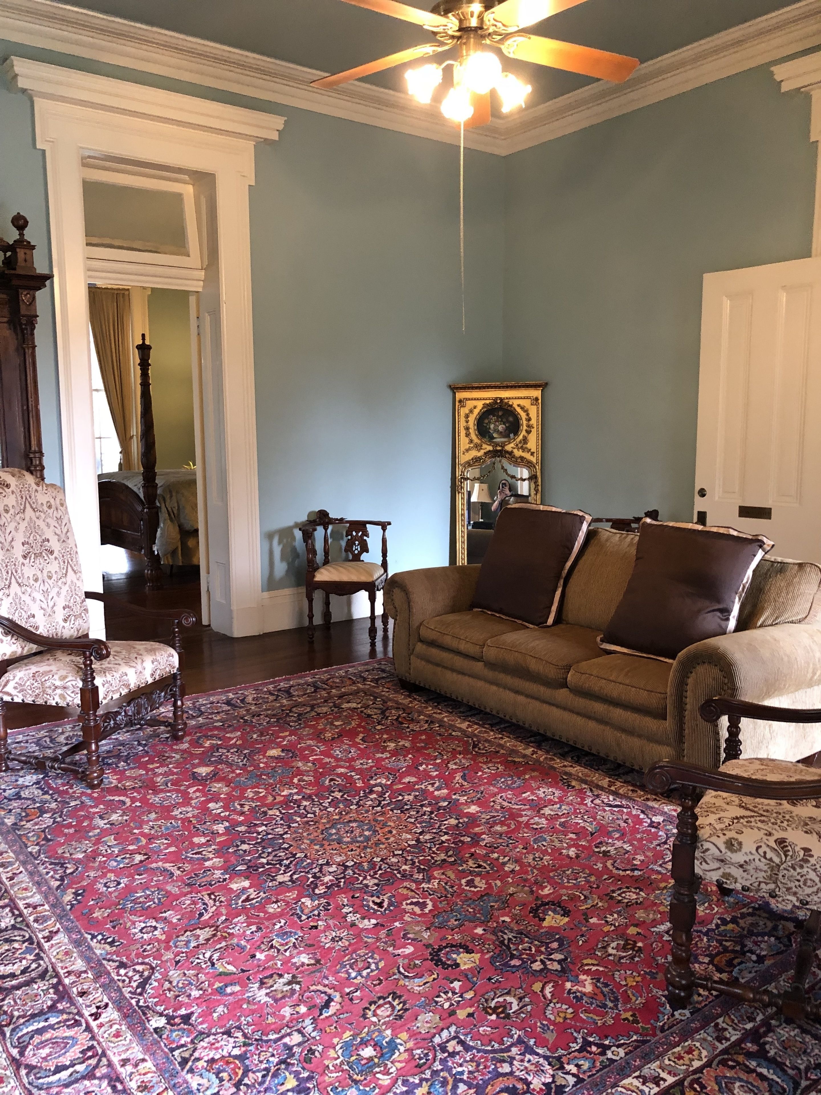 Een woonkamer met een kamerkleed met patroon, een bruine bank, een houten stoel en een plafondventilator.