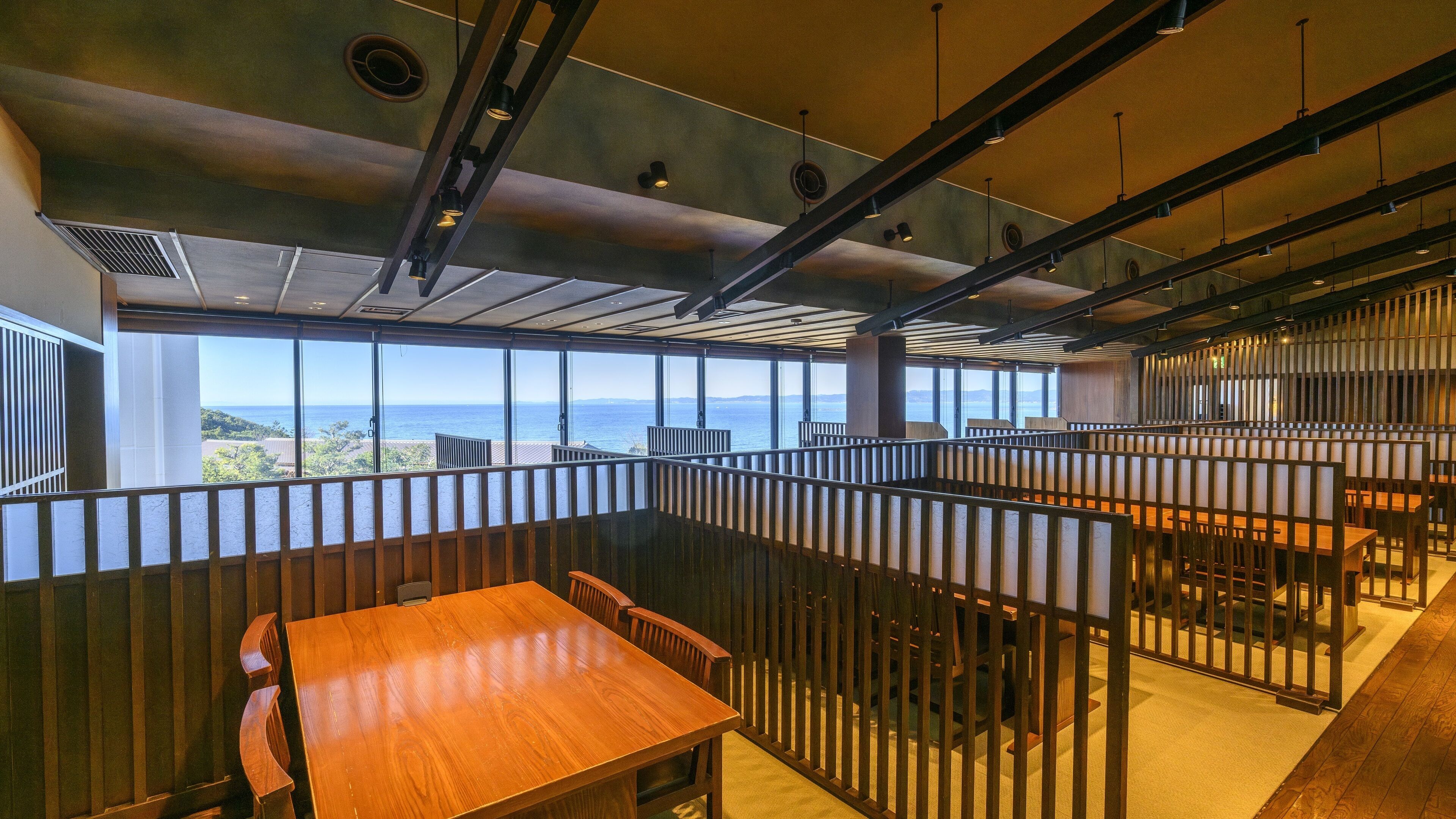 A modern restaurant with high ceilings, large windows offering ocean views, and a mix of wooden and metal furnishings.