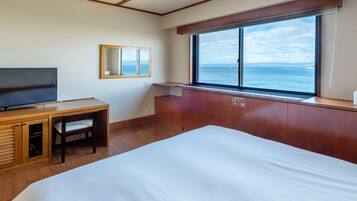 A hotel room with a bed, a desk with a chair, a television, and a large window with a view of the sea.