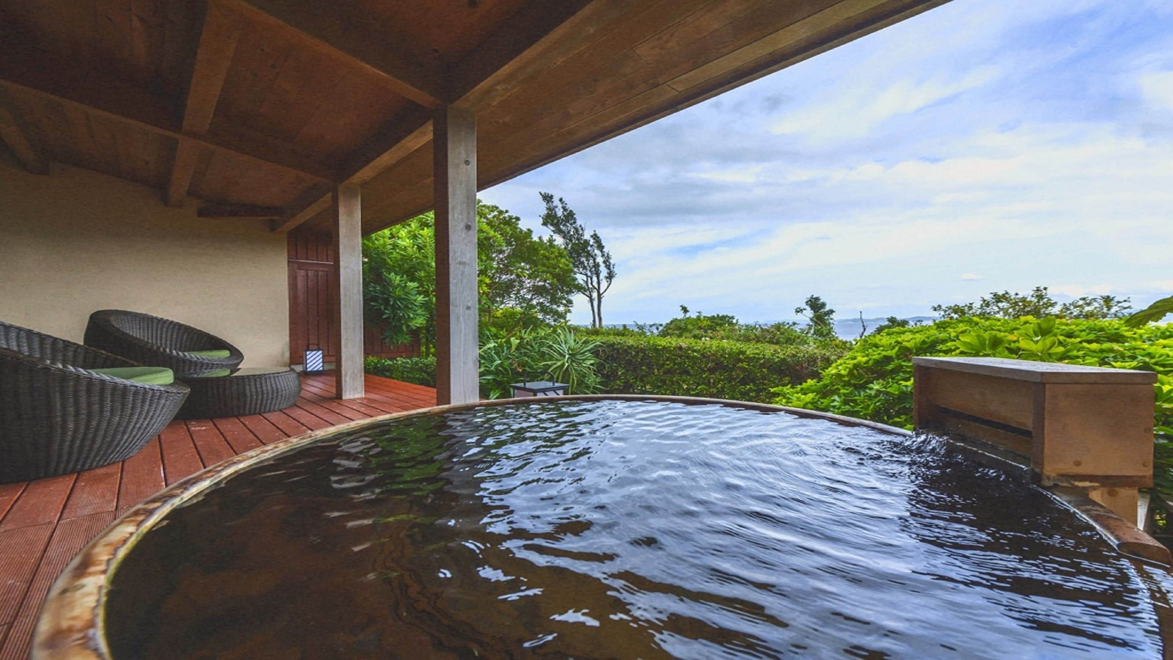 A hot tub with a wooden deck and wicker furniture, surrounded by lush greenery and a view of the sea.