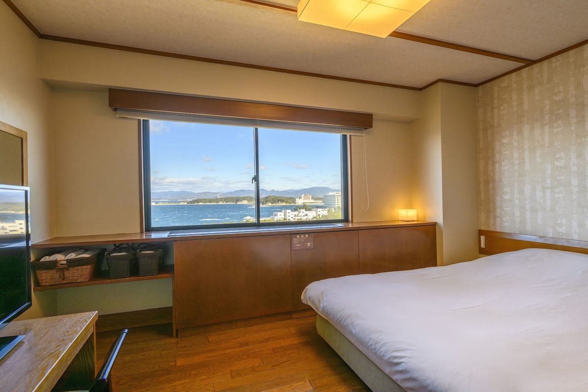 Een hotelkamer met een bed, een bureau met een televisie, een raam met uitzicht op zee en gebouwen, en een nachtkastje met een lamp.