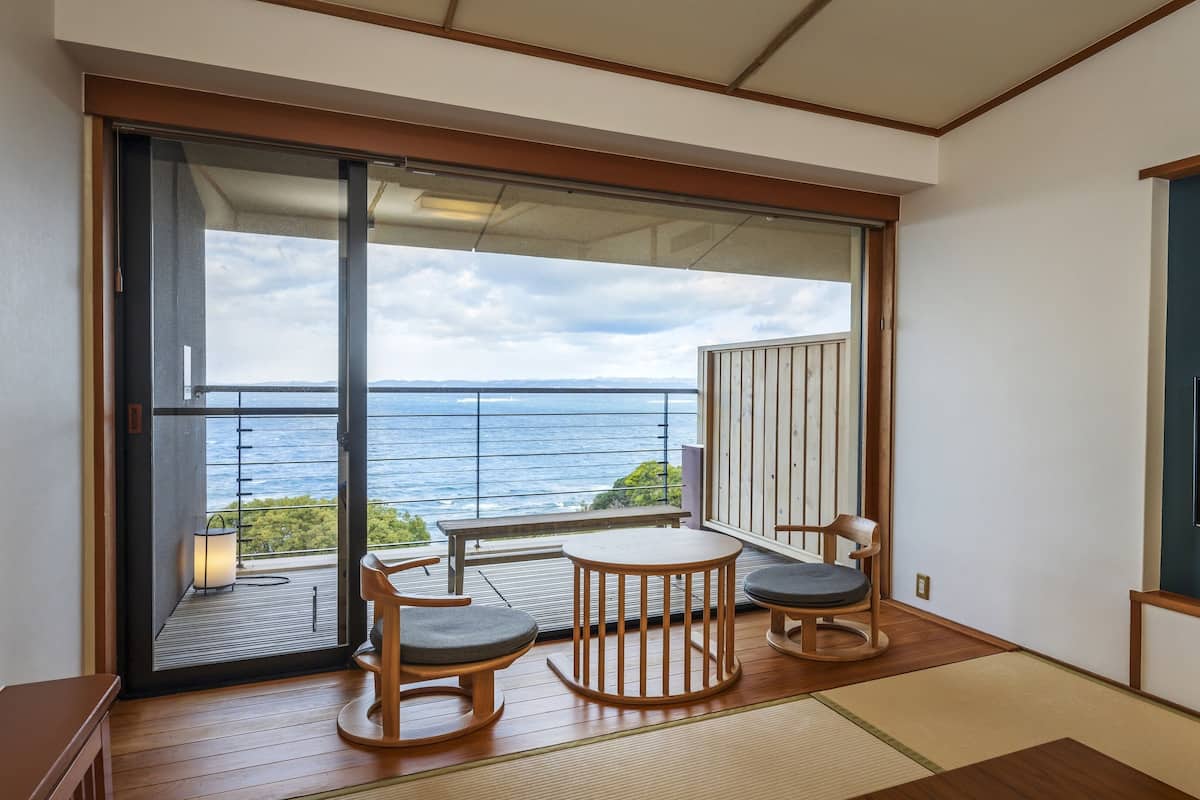 Een kamer met een schuifpui die uitkomt op een balkon met uitzicht op zee. Er staat een klein, rond tafel met twee stoelen en een aan de muur bevestigde televisie.