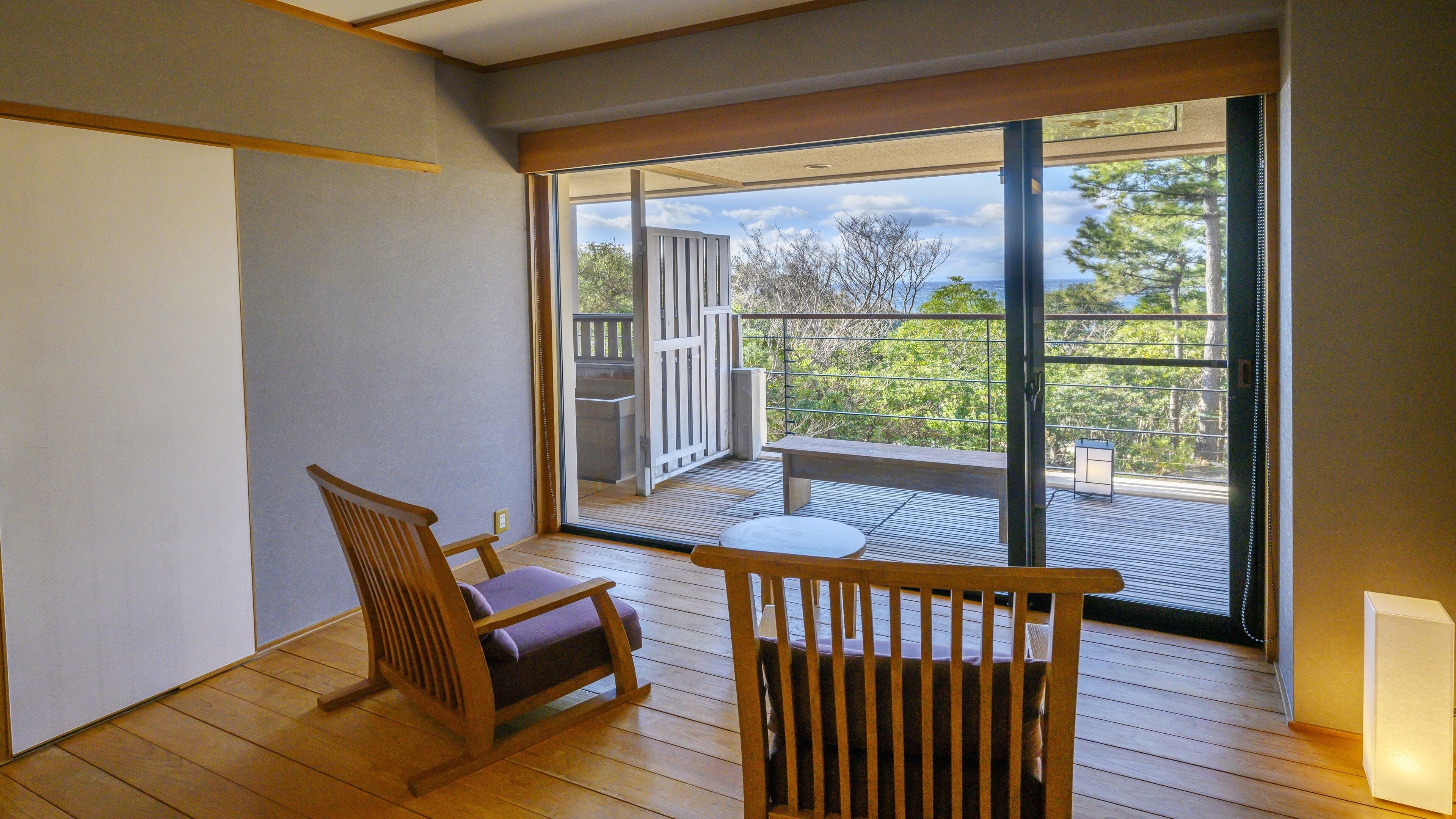 Habitación con piso de madera, una silla de madera, una pequeña mesa y un ventanal corredero que da a un balcón exterior con vistas a árboles y barandilla.