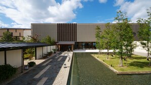 Een modern gebouw met een plat dak, een overdekte doorgang en een kleine tuin met bomen en een waterpartij.