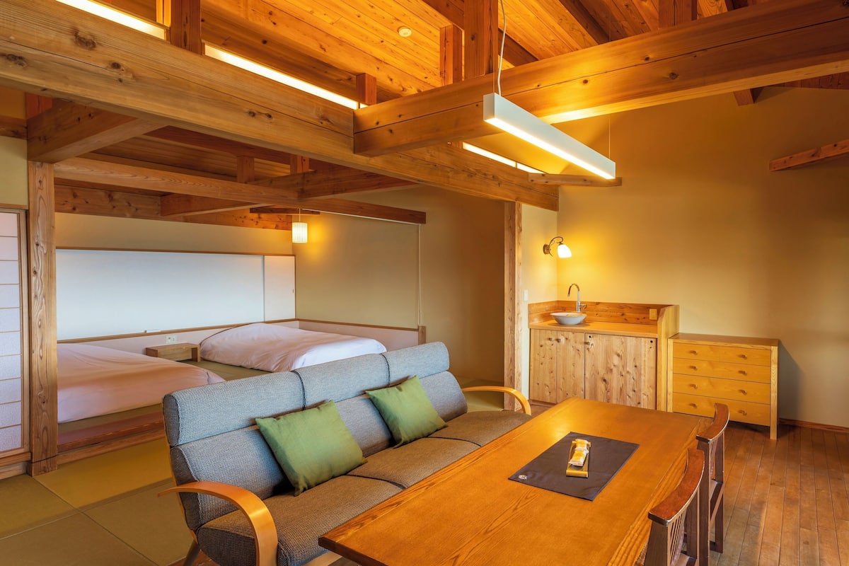 Een knus ingerichte kamer met een houten plafond, een bed, een bank met kussens, een houten tafel en een klein houten kastje met een wastafel.