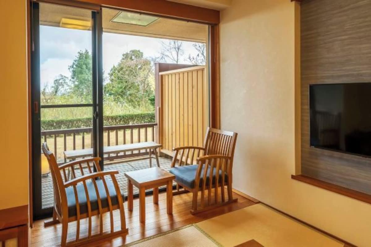 Una habitación con puerta corrediza de vidrio que da a un balcón, una mesa de madera con sillas y un televisor montado en la pared.