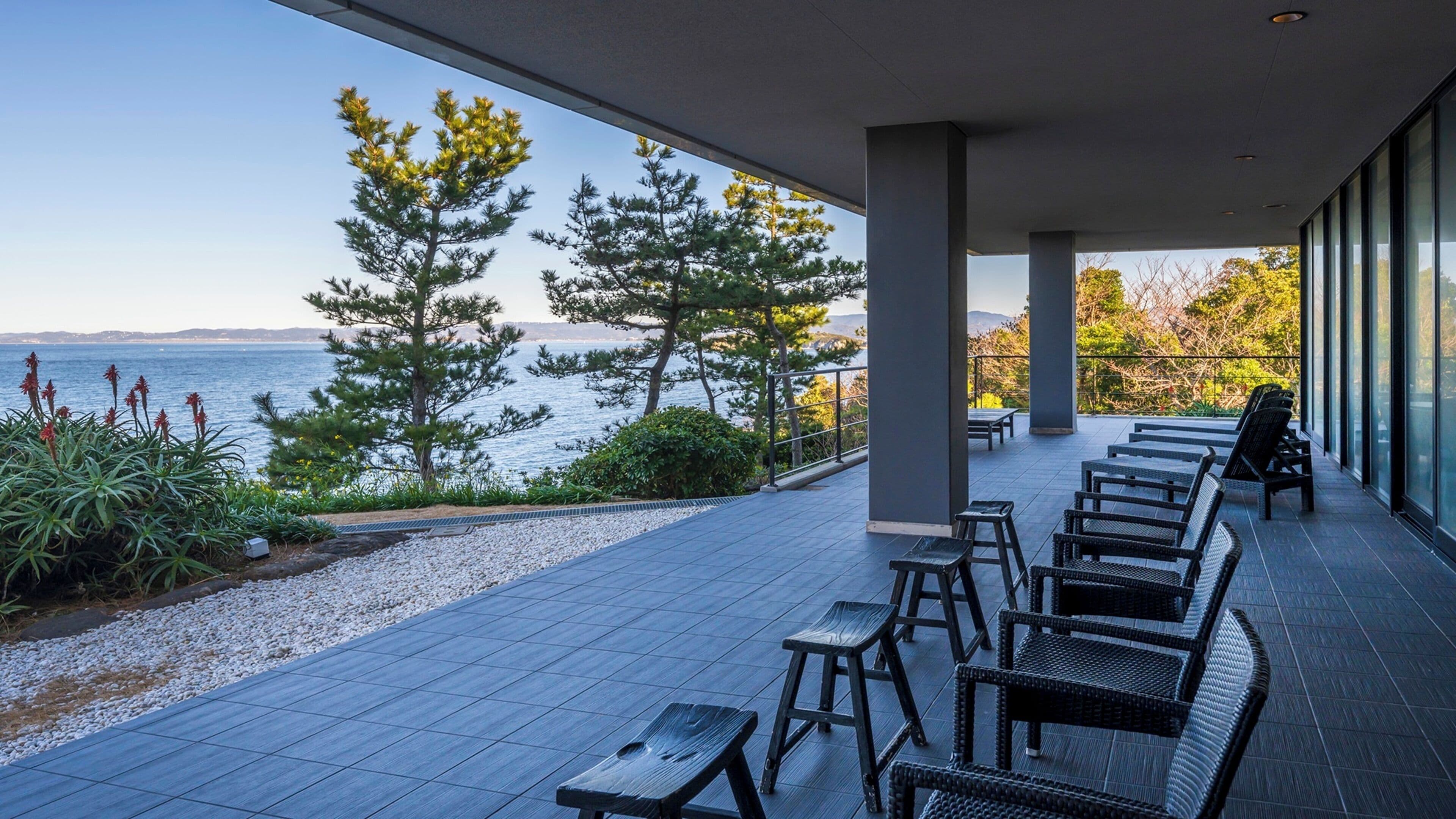 A covered outdoor seating area with tables and chairs, overlooking a body of water and trees.