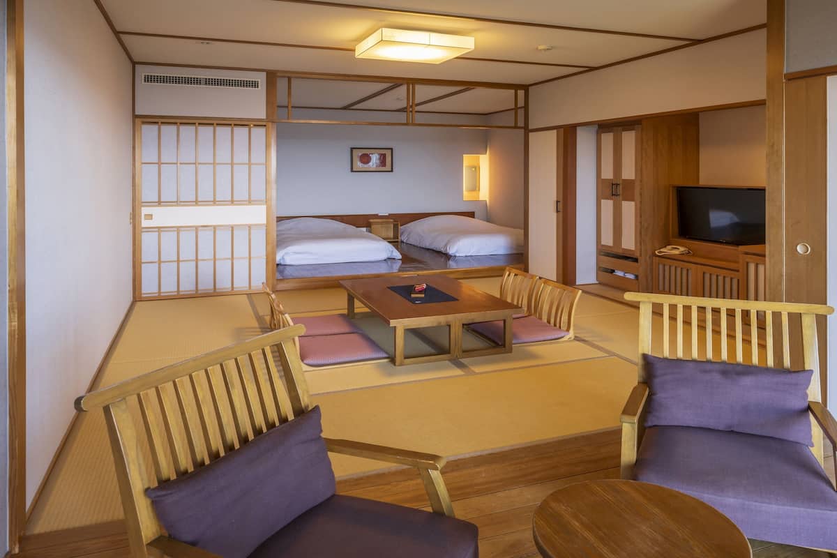 Een traditionele Japanse kamer met tatami vloeren, een lage tafel en twee tatami matten met kussens. Er staat een bed met wit beddengoed en een televisie in de hoek.