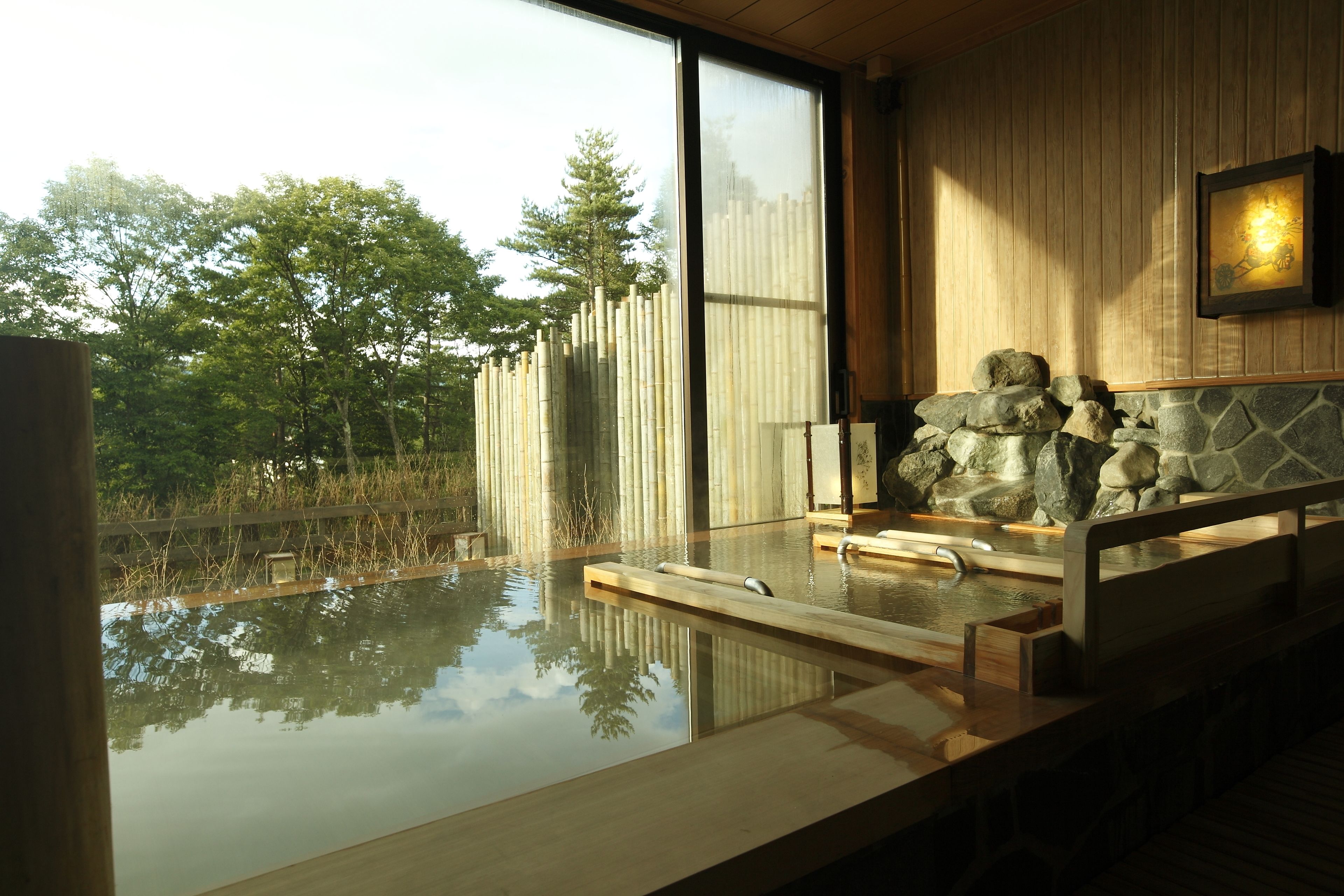 A serene indoor hot spring area with a wooden deck, a stone wall, and a view of trees and bamboo outside.
