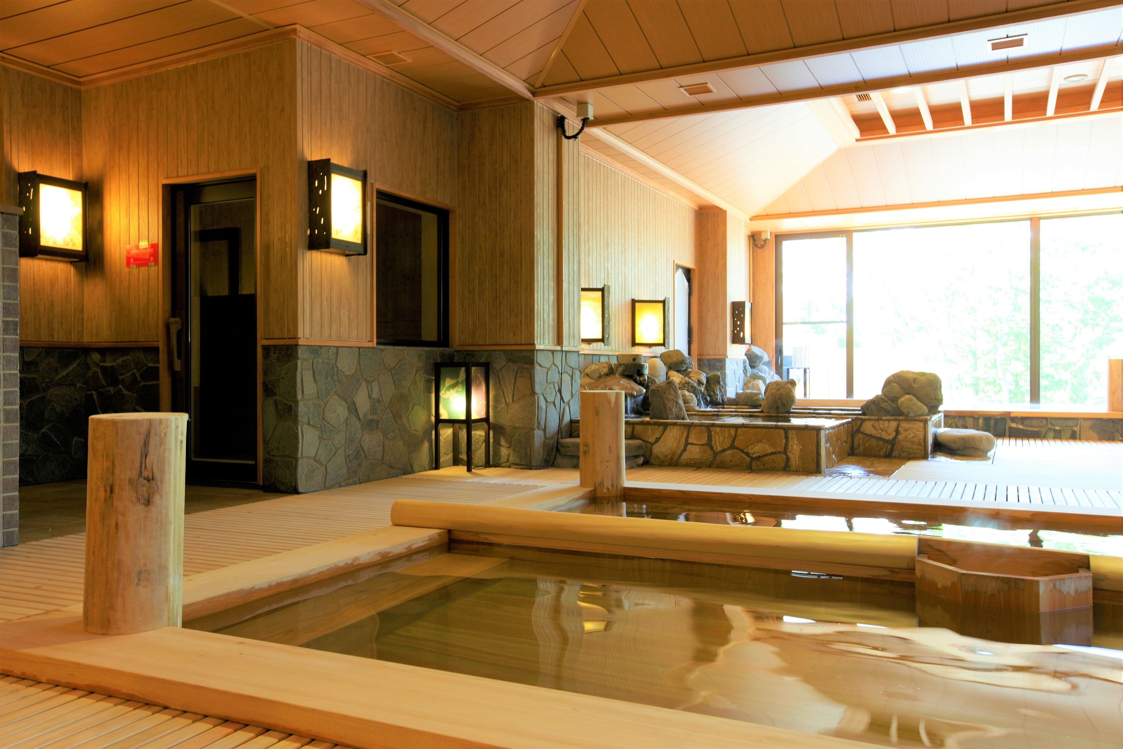 Un onsen japonés tradicional con un baño de piedra, bancos de madera y vistas al exterior a través de amplios ventanales.