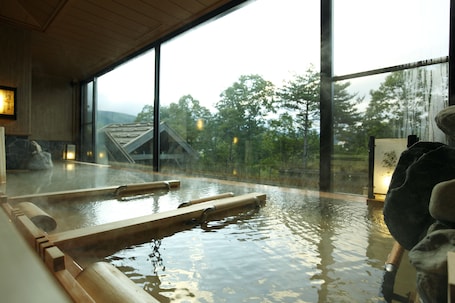 A hot spring bath with wooden benches, a stone wall, and a view of trees through large windows.