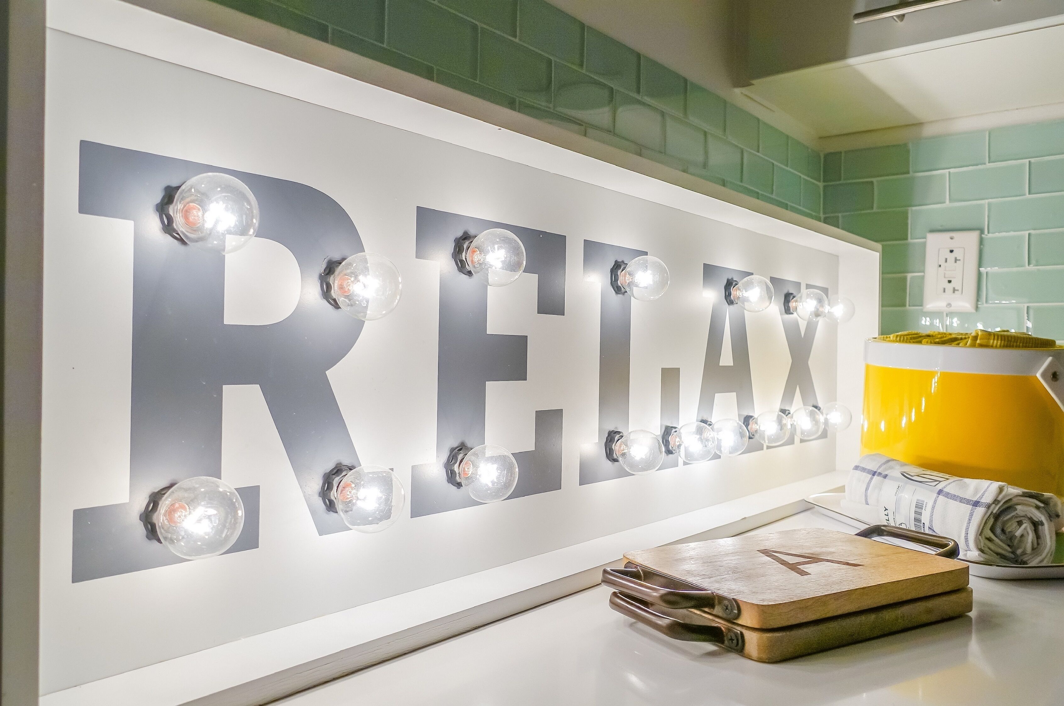 A wall with the word "RELAX" illuminated by lights, a yellow lamp, and a wooden cutting board with a letter 'A' on it.