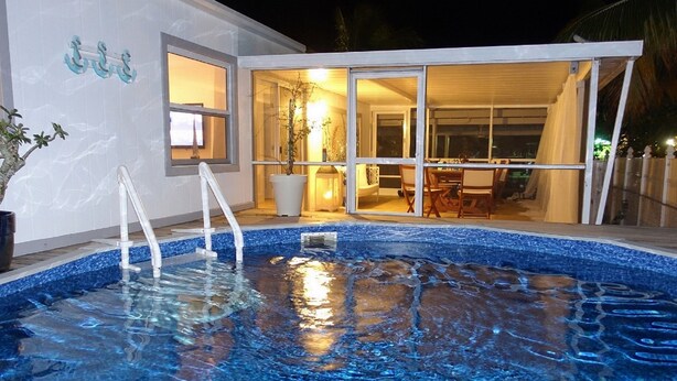 A swimming pool with clear blue water, a white exterior wall, and a glass door leading to an indoor area with a table and chairs. - Beautiful Waterfront Villa (Tavernier)