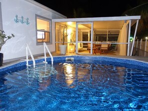 A swimming pool with clear blue water, a white exterior wall, and a glass door leading to an indoor area with a table and chairs.