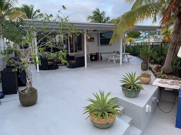 A covered outdoor seating area with a patio table and chairs, surrounded by potted plants and a palm tree.