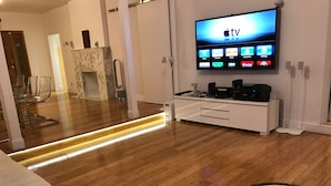 A modern living room with a wooden floor, a flat-screen TV mounted on the wall displaying various streaming services, a white entertainment unit with electronic devices, and a patterned rug. - 3Bd. & 2Bth. Private Island Security Gated Villa Miami Beach (North Bay Village)