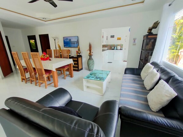 A living room with a black leather sofa, a dining table with chairs, a TV, and a kitchen area in the background.