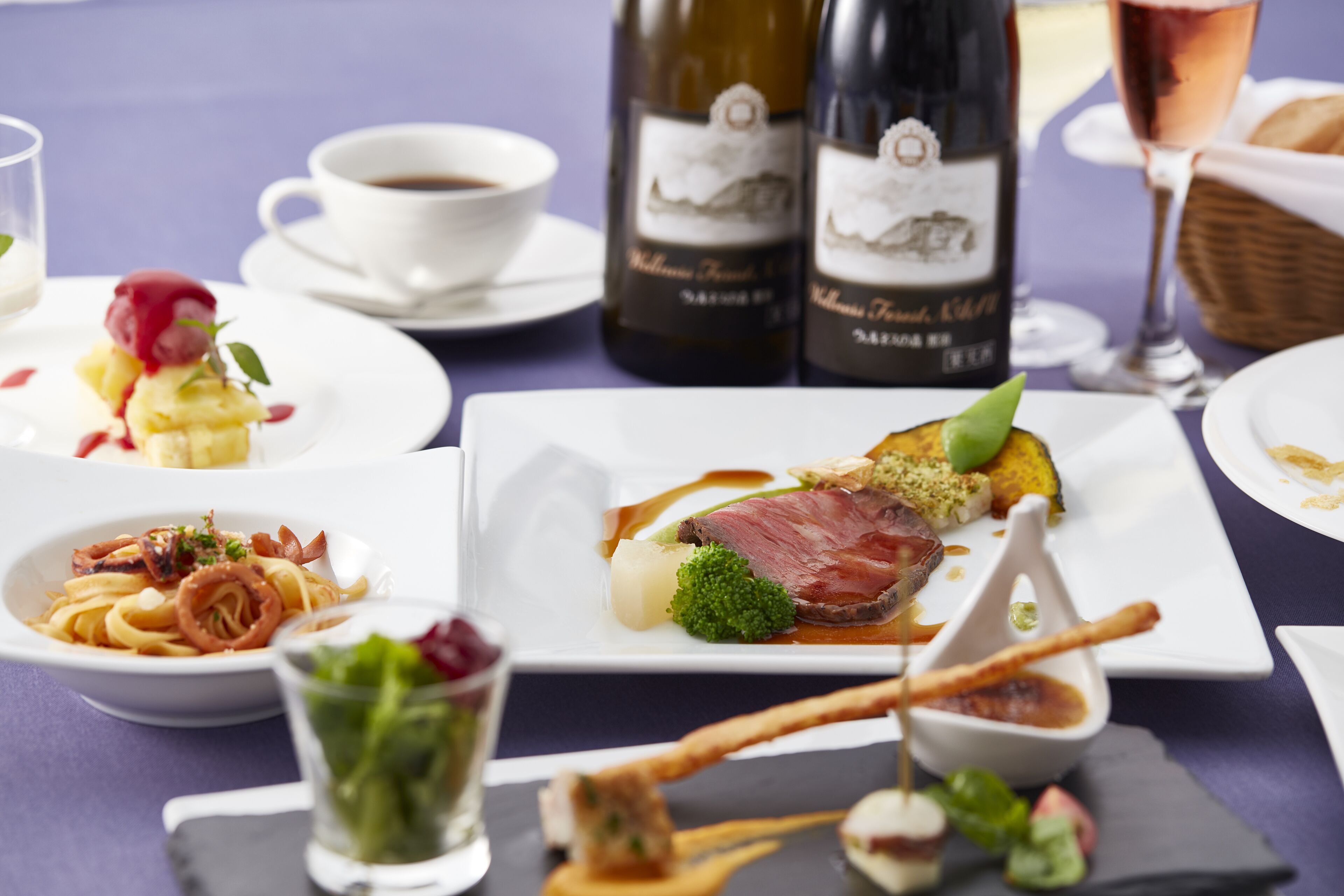 Uma mesa posta com diversos pratos, incluindo massa, bife e legumes, além de garrafas de vinho e copos.