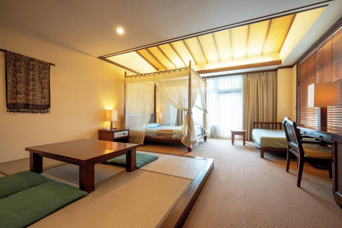 A traditional Japanese-style room with tatami flooring, a low wooden table, a futon bed, a desk with a chair, and a canopy bed.